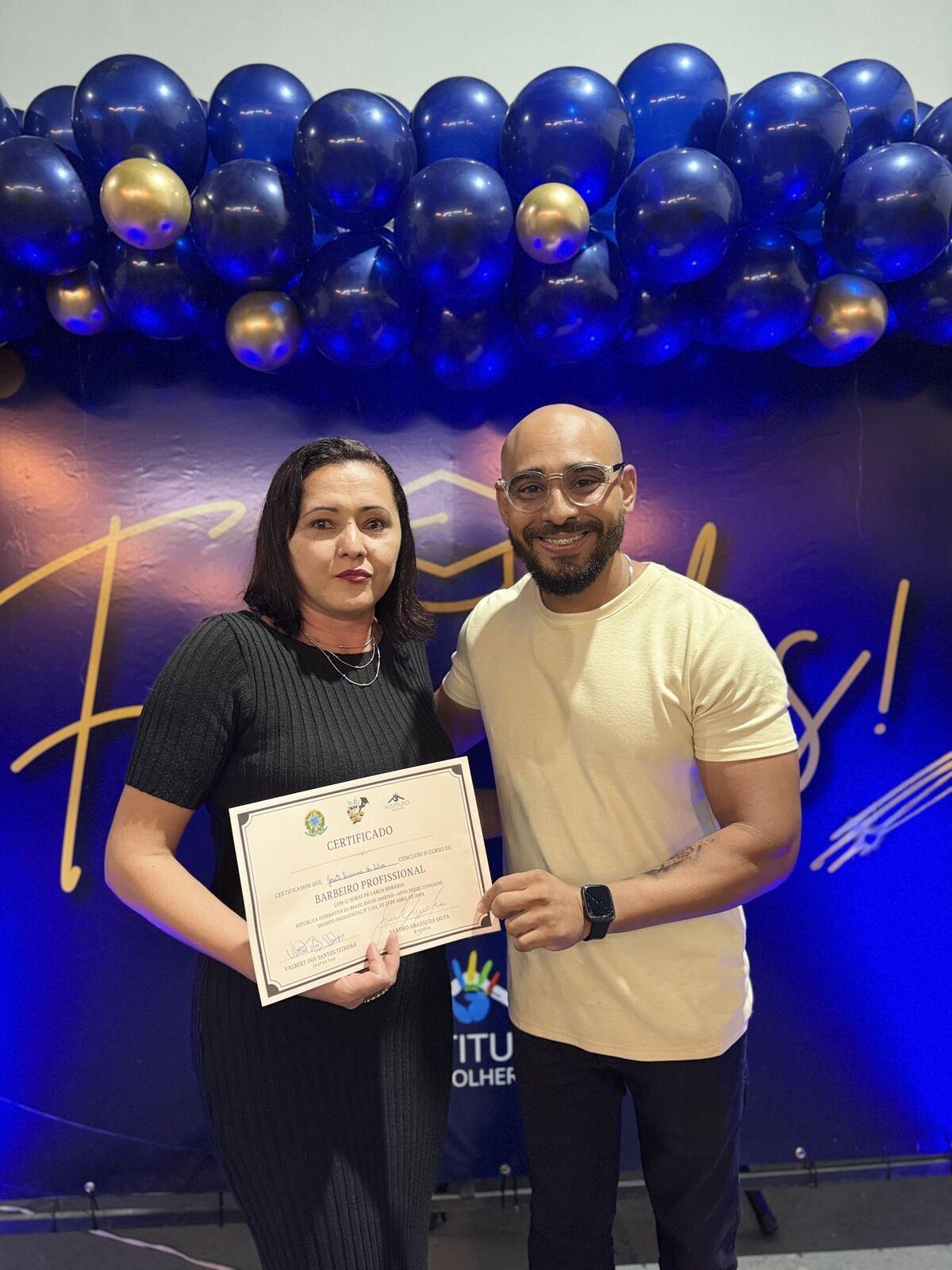 Formanda do curso de Barbeiro e o professor, juntos, segurando o certificado diante do painel de balões azuis da formatura