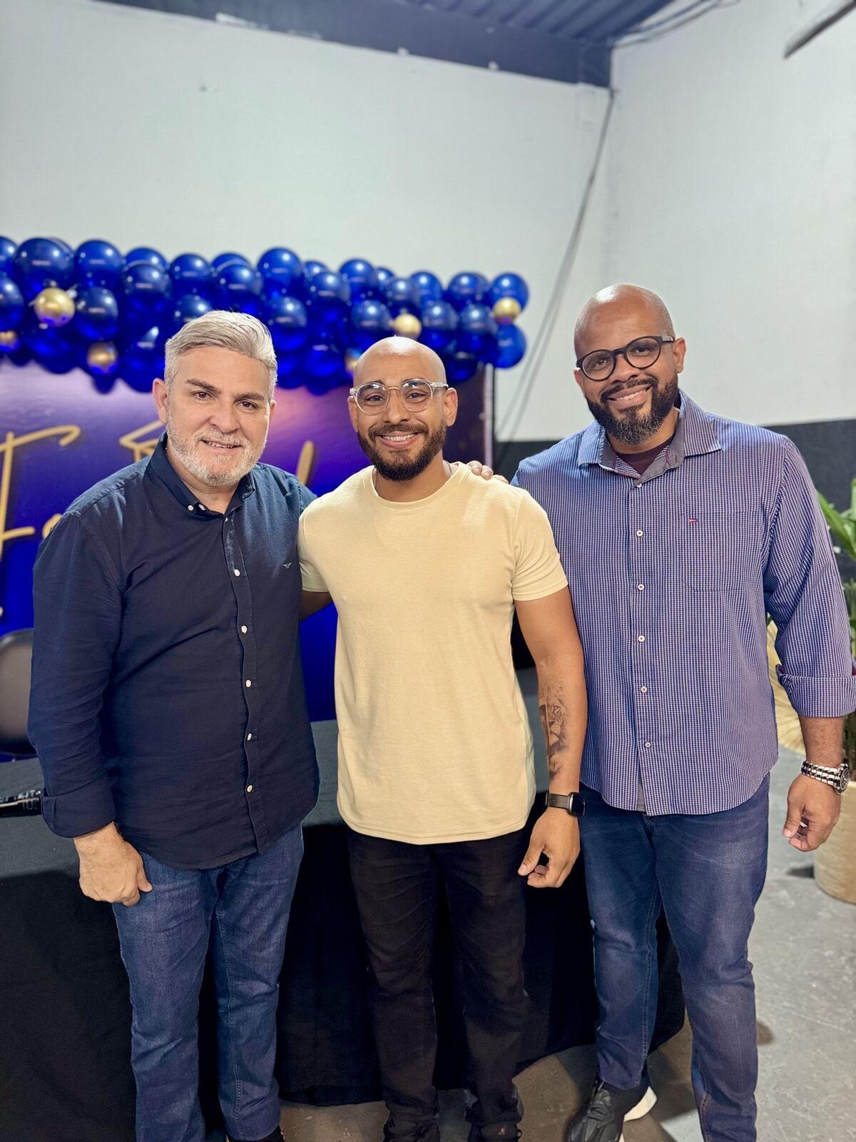 Professor do curso de Barbeiro com dois convidados na formatura, sob o painel de balões azuis