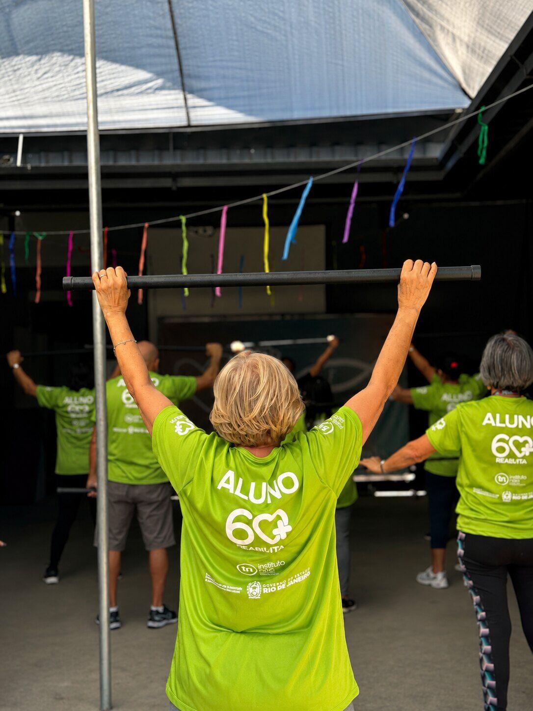 Alunos de camiseta verde fazendo exercício com barra, vistos de costas, sob bandeirinhas coloridas