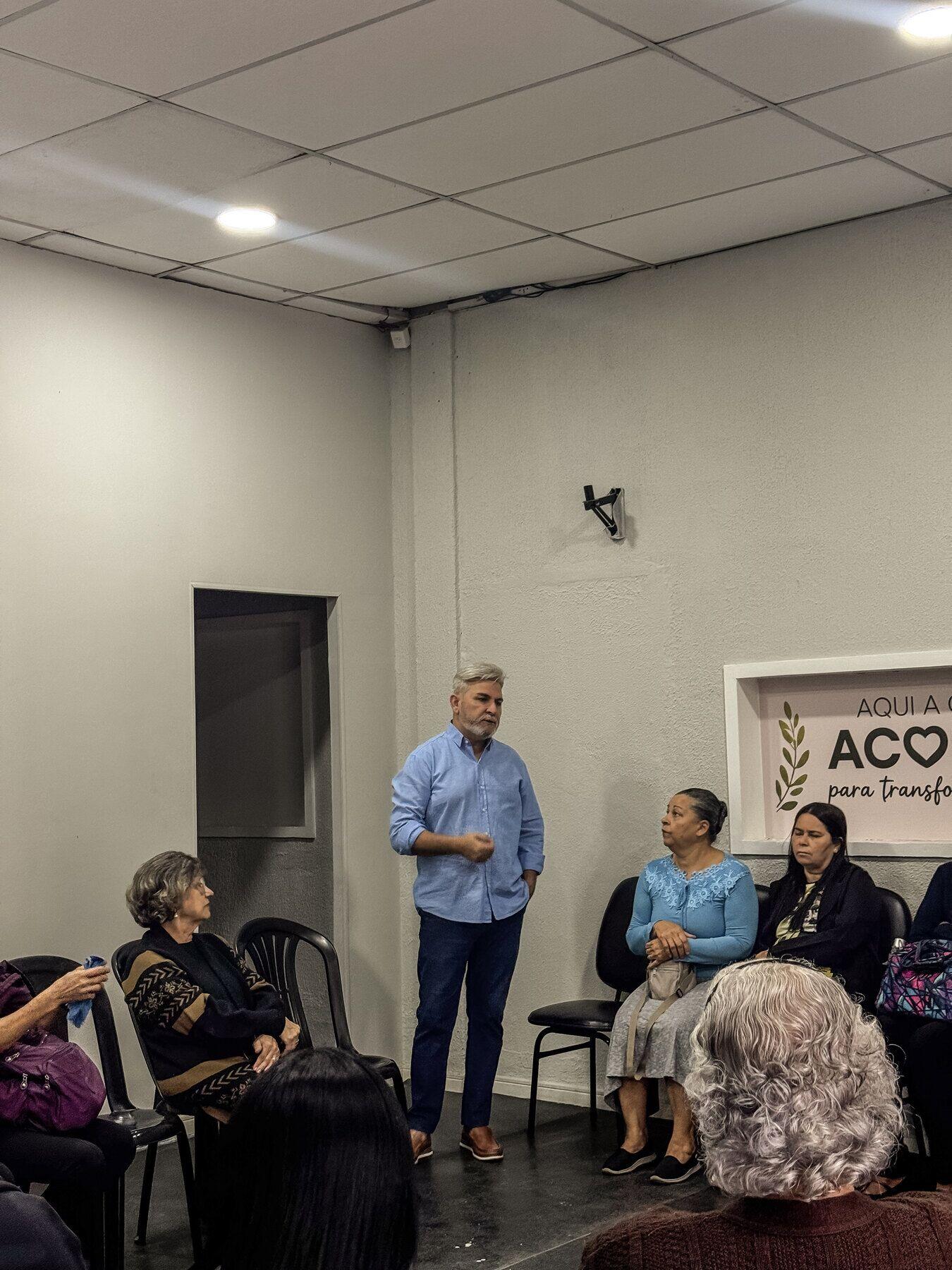 Homem de camisa azul falando para o grupo, ao lado do letreiro “Aqui a gente acolhe”